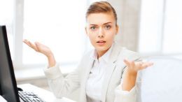 Business woman sitting at desk with palms facing up asking, "how what?"
