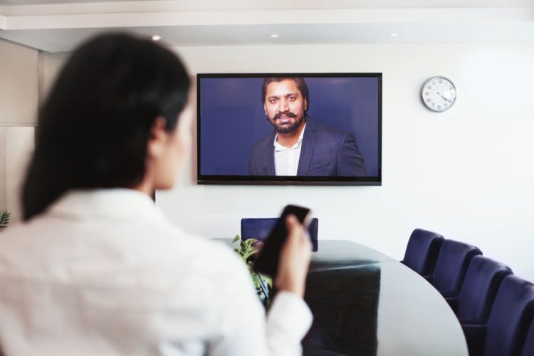 A senior executive appears on the TV screen in a conference room before a virtual meeting.